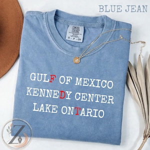 Op de afbeelding: Een spijkerblauw t-shirt met korte mouwen en een ronde hals. Het shirt heeft witte tekst met de woorden GULF OF MEXICO, KENNEDY CENTER en LAKE ONTARIO, waarbij de letters F, D en T in het rood zijn gemarkeerd. Een gouden ketting ligt erbovenop.