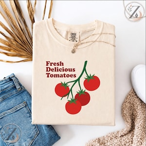 Può includere: Una t-shirt color crema con una grafica di quattro pomodori rossi su un ramo verde. Il testo "Fresh Delicious Tomatoes" è stampato in rosso a sinistra dei pomodori. La maglietta presenta un'etichetta del marchio Comfort Colors sul collo.
