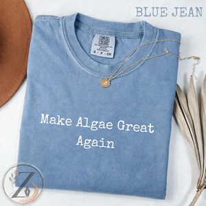 Könnte beinhalten: Ein kurzärmeliges T-Shirt in Jeansblau mit Rundhalsausschnitt. Das T-Shirt ist auf der Vorderseite mit dem weißen Schriftzug "Make Algae Great Again" bedruckt. Eine goldfarbene Kette mit einem kleinen runden Anhänger liegt am Kragen.