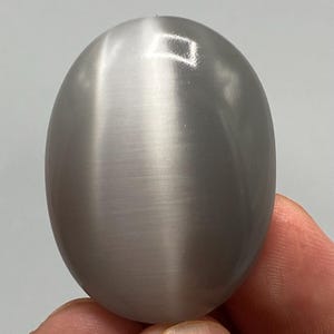 May include: An oval-shaped, smooth, gray gemstone featuring a distinct chatoyant band of light running vertically across its center, creating a cat's eye effect with a polished, reflective surface that catches the light.
