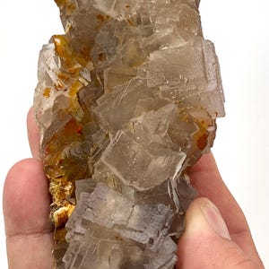Op de afbeelding: Een cluster van doorschijnende, kubusvormige fluorietkristallen met een lichtgrijze en rookbruine kleur. Het mineraalexemplaar vertoont scherpe, geometrische randen en gelaagde kristalstructuren met zichtbare oranjebruine mineraalafzettingen.