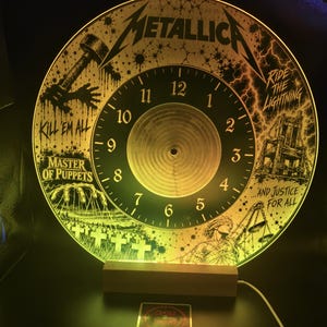 Puede incluir: Un reloj de pared circular con un diseño gráfico de Metallica que incluye portadas de los álbumes Kill 'Em All, Ride the Lightning, Master of Puppets y ...And Justice for All. La esfera está iluminada con luz amarilla cálida sobre una base de madera.
