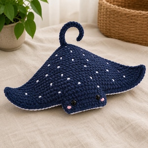 May include: A dark navy blue crochet stingray plush toy featuring a white speckled pattern on its back, a curved tail, two black plastic eyes, and small pink embroidered cheeks. The underside of the stingray is white, and the texture is yarn.
