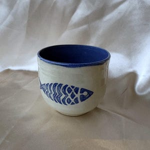 Könnte beinhalten: Ein Keramikbecher mit cremefarbener Außenseite und tiefblauem Innenraum. Die Außenfläche ist mit einem handgemalten blauen Fischskelett-Motiv verziert. Der Becher hat eine abgerundete, henkellose Form mit einer glatten Oberfläche und einem Rand.