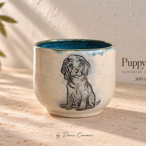 Puede incluir: Una taza de cerámica color crema con interior azul, que presenta una ilustración de un cachorro dibujada a mano en el frente. El texto Puppy love, HANDMADE CERAMICS, 300 ml y by Dione Ceramic está impreso en y cerca de la taza.