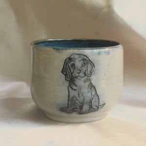 Peut inclure: Une tasse en céramique avec un vernis blanc cassé brillant et un bord teinté de bleu. L'extérieur présente une illustration dessinée à la main d'un chiot à l'encre gris foncé. La tasse a une forme arrondie sans anse avec une finition lisse.