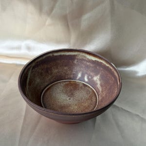 Pode incluir: Uma tigela de cerâmica redonda com um exterior texturizado castanho escuro e um interior salpicado em tons de bege claro e castanho. A tigela apresenta uma borda larga e aberta com uma base lisa e curva, exibindo um acabamento natural.