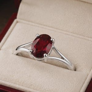 K&ouml;nnte beinhalten: Ein silberfarbener Ring mit einem gro&szlig;en, oval geschliffenen, tiefroten Edelstein in einer Vier-Krappen-Fassung. Die Ringschiene ist gespalten und l&auml;uft zur Mitte hin schmaler zu, was eine polierte metallische Oberfl&auml;che und einen facettierten Stein zeigt.