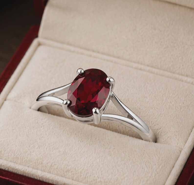 K&ouml;nnte beinhalten: Ein silberfarbener Ring mit einem gro&szlig;en, oval geschliffenen, tiefroten Edelstein in einer Vier-Krappen-Fassung. Die Ringschiene ist gespalten und l&auml;uft zur Mitte hin schmaler zu, was eine polierte metallische Oberfl&auml;che und einen facettierten Stein zeigt.