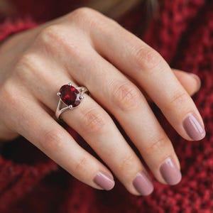 K&ouml;nnte beinhalten: Ein silberner Ring mit einem gro&szlig;en, oval geschliffenen, tiefroten Edelstein in einer Vier-Krappen-Fassung. Die Ringschiene weist ein geteiltes Design auf, das sich zur Mitte hin verj&uuml;ngt und eine polierte Metalloberfl&auml;che zeigt, die den Stein betont.