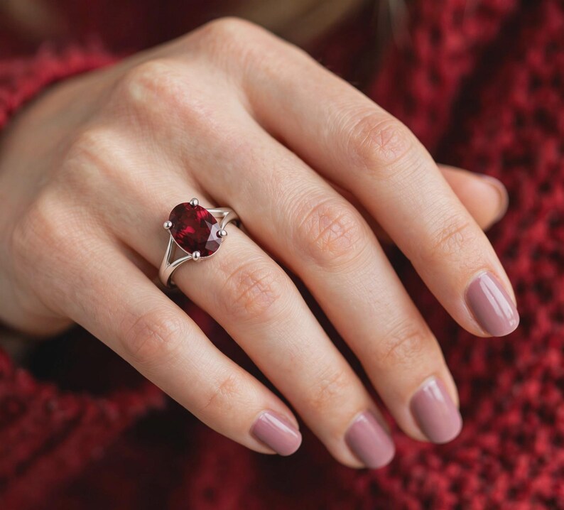 K&ouml;nnte beinhalten: Ein silberner Ring mit einem gro&szlig;en, oval geschliffenen, tiefroten Edelstein in einer Vier-Krappen-Fassung. Die Ringschiene weist ein geteiltes Design auf, das sich zur Mitte hin verj&uuml;ngt und eine polierte Metalloberfl&auml;che zeigt, die den Stein betont.