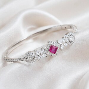 Può includere: Un bracciale rigido incernierato color argento con una pietra preziosa centrale di taglio quadrato color rosa vivace circondata da un alone di piccole pietre chiare. Il design include accenti floreali con pietre chiare taglio marquise.