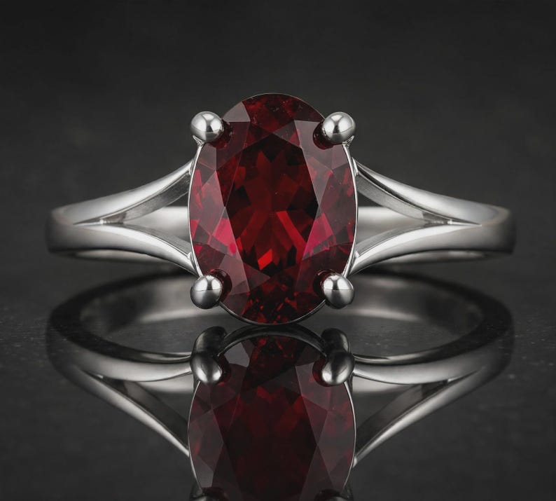 K&ouml;nnte beinhalten: Ein silberfarbener Ring mit einem gro&szlig;en, oval geschliffenen, tiefroten Edelstein, der von vier Krappen gehalten wird. Die Ringschiene hat ein geteiltes Design, das sich zur Mitte hin verj&uuml;ngt und f&uuml;r ein poliertes, reflektierendes Aussehen sorgt.