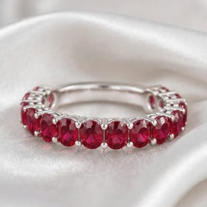 Könnte beinhalten: Ein silberner Eternity-Ring mit einer durchgehenden Reihe oval geschliffener, leuchtend roter Rubine, die in polierten Metallfassungen sitzen. Der Ring zeigt eine gleichmäßige Anordnung von Edelsteinen rund um das Band in einem satten Karmesinrot.