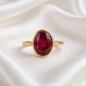 Puede incluir: Un anillo de oro con una gran piedra preciosa de color rojo intenso y corte ovalado, engastada en un bisel de oro liso y pulido. El aro es fino y circular, fabricado en metal dorado a juego, con un diseño limpio y clásico de piedra facetada.
