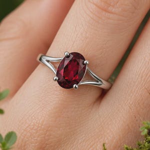 K&ouml;nnte beinhalten: Ein silberfarbener Ring mit einem zentralen, oval geschliffenen, tiefroten Edelstein, der von vier Krappen gehalten wird. Die Ringschiene ist gespalten und verj&uuml;ngt sich zur Fassung hin, was f&uuml;r ein glattes und poliertes Aussehen sorgt.