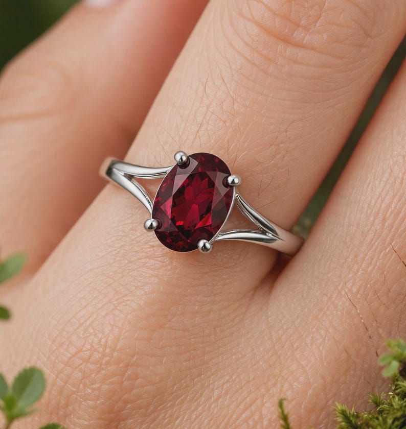 K&ouml;nnte beinhalten: Ein silberfarbener Ring mit einem zentralen, oval geschliffenen, tiefroten Edelstein, der von vier Krappen gehalten wird. Die Ringschiene ist gespalten und verj&uuml;ngt sich zur Fassung hin, was f&uuml;r ein glattes und poliertes Aussehen sorgt.