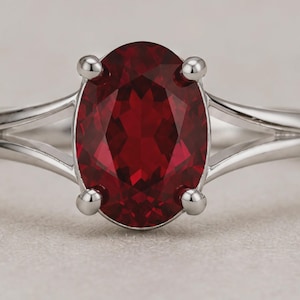 K&ouml;nnte beinhalten: Ein Silberring mit einem gro&szlig;en, oval geschliffenen, tiefroten Edelstein, der von vier polierten Krappen gehalten wird. Die Ringschiene hat ein geteiltes Design, das sich zur zentralen Fassung hin verj&uuml;ngt und ein glattes Metallfinish zeigt.