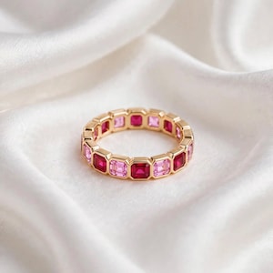 Könnte beinhalten: Ein goldener Eternity-Ring mit einer durchgehenden Reihe abwechselnd quadratisch geschliffener rosa und tiefroter Edelsteine. Die Steine sind in einen polierten Goldrahmen gefasst, was ein gleichmäßiges, kreisförmiges Design mit Farbverlauf ergibt.