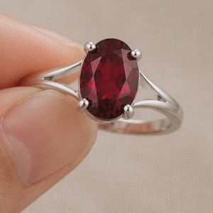 K&ouml;nnte beinhalten: Ein silberfarbener Ring mit einem gro&szlig;en, oval geschliffenen, tiefroten Edelstein in einer Vier-Krappen-Fassung. Die Ringschiene ist gespalten und verj&uuml;ngt sich zum Mittelstein hin, was dem Schmuckst&uuml;ck ein glattes und poliertes Aussehen verleiht.