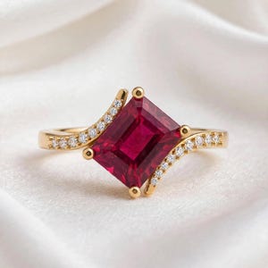 Könnte beinhalten: Ein Goldring mit einem großen, rechteckigen, tiefroten Edelstein, der schräg eingesetzt ist. Die Ringschiene teilt sich in zwei asymmetrische, geschwungene Arme, die den Mittelstein umfassen und mit kleinen, klaren Diamanten besetzt sind.