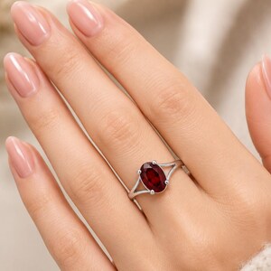 K&ouml;nnte beinhalten: Ein silberner Ring mit einem oval geschliffenen, tiefroten Granat-Edelstein, der von vier Krappen gehalten wird. Die Ringschiene hat ein geteiltes Design, das sich zur Mitte hin verj&uuml;ngt und ein poliertes Finish mit dem dunklen Edelstein bildet.