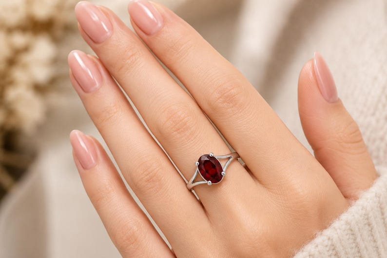 K&ouml;nnte beinhalten: Ein silberner Ring mit einem oval geschliffenen, tiefroten Granat-Edelstein, der von vier Krappen gehalten wird. Die Ringschiene hat ein geteiltes Design, das sich zur Mitte hin verj&uuml;ngt und ein poliertes Finish mit dem dunklen Edelstein bildet.