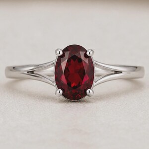 K&ouml;nnte beinhalten: Ein silberfarbener Ring mit einem zentralen, oval geschliffenen, tiefroten Edelstein, der von vier Krappen gehalten wird. Die Ringschiene hat ein geteiltes Design, das sich zur Fassung hin verj&uuml;ngt und dem Schmuckst&uuml;ck ein poliertes Aussehen verleiht.