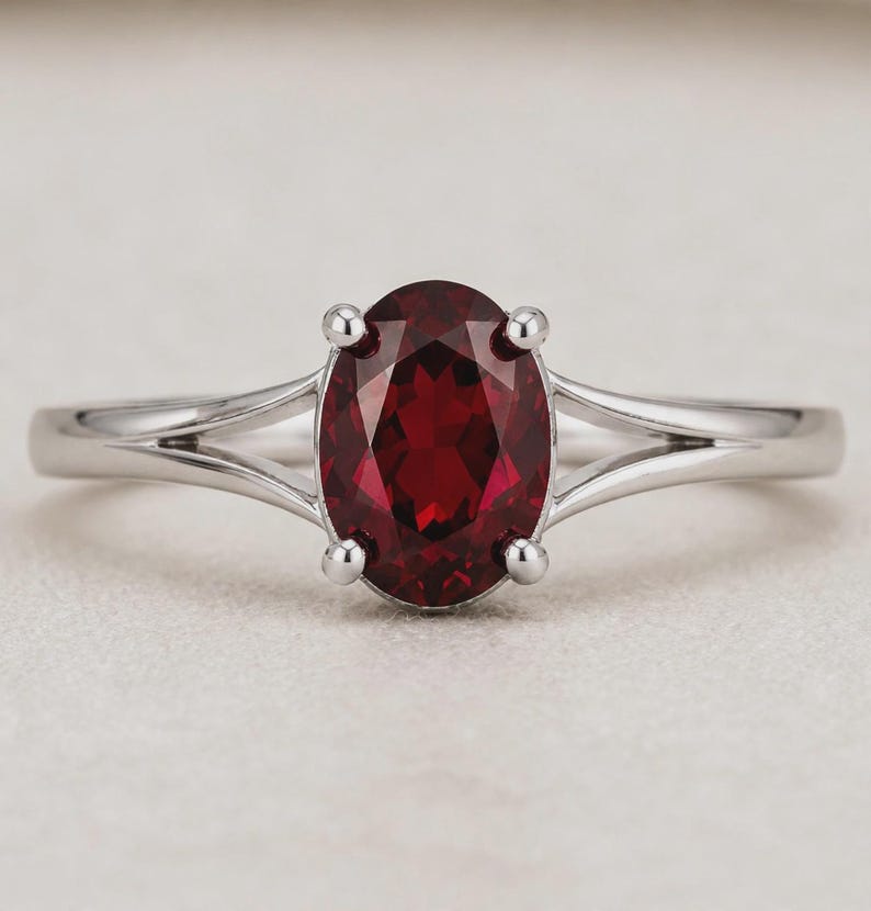 K&ouml;nnte beinhalten: Ein silberfarbener Ring mit einem zentralen, oval geschliffenen, tiefroten Edelstein, der von vier Krappen gehalten wird. Die Ringschiene hat ein geteiltes Design, das sich zur Fassung hin verj&uuml;ngt und dem Schmuckst&uuml;ck ein poliertes Aussehen verleiht.
