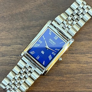 Könnte beinhalten: Eine rechteckige Seiko Quartz Armbanduhr mit tiefblauem Zifferblatt, silberfarbenen römischen Ziffern und silbernen Zeigern. Die Uhr verfügt über ein poliertes, silberfarbenes Metallgliederarmband mit mehrreihigem Design und eine kleine Krone.