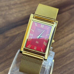 May include: A rectangular gold-tone wristwatch featuring a vibrant red dial with a textured pattern. The watch face displays gold Roman numeral hour markers, gold hands, and the text Seiko Quartz. It includes a gold-tone metal mesh strap.