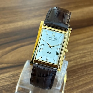 May include: A rectangular Seiko Quartz watch with a gold-tone case and a white dial featuring black Roman numeral hour markers. The watch has a dark brown leather strap with a textured crocodile-embossed pattern and a gold-tone crown on the side.