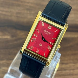 Peut inclure: Une montre-bracelet rectangulaire Seiko Quartz dotée d'un cadran rouge vif avec des index en chiffres romains dorés et des aiguilles noires. La montre possède un boîtier doré poli et un bracelet en cuir noir à la finition texturée.