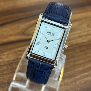 May include: A rectangular Seiko Quartz wristwatch with a silver-tone case and a white dial featuring black Roman numeral hour markers. The watch has a textured blue leather strap with a crocodile-embossed pattern and a small crown on the side.