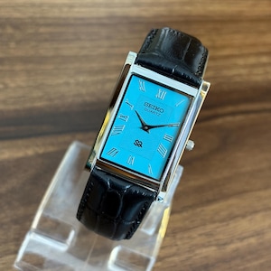 May include: A rectangular Seiko Quartz wristwatch featuring a vibrant turquoise dial with black Roman numeral hour markers and black hands. The watch has a polished silver-tone metal case and a black leather strap with a textured crocodile pattern.