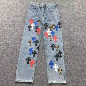 Pode incluir: Calças de ganga azul claro com cintura subida e pernas direitas. A parte da frente das calças está decorada com vários emblemas em forma de cruz em diversas cores, incluindo vermelho, azul, branco, preto e um padrão de leopardo.