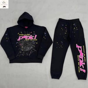 May include: A black hooded sweatshirt and matching sweatpants set. The hoodie features a white spiderweb graphic, pink text reading "pink!", and small colourful stars. The trousers include the same pink text and scattered colourful star accents.