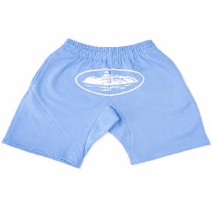 May include: Light blue shorts featuring an elastic waistband and a white graphic print centred on the back. The graphic depicts a lighthouse on a rocky island surrounded by water, enclosed within a white oval outline on the fabric.
