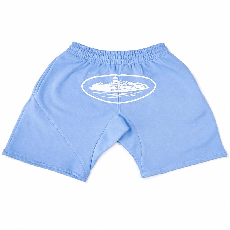 May include: Light blue shorts featuring an elastic waistband and a white graphic print centred on the back. The graphic depicts a lighthouse on a rocky island surrounded by water, enclosed within a white oval outline on the fabric.