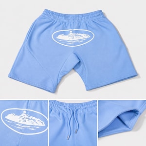 May include: Light blue athletic shorts featuring an elastic waistband with a drawstring and a white oval graphic on the front depicting a lighthouse on a rocky island. The shorts have a soft interior lining and visible side seam construction.