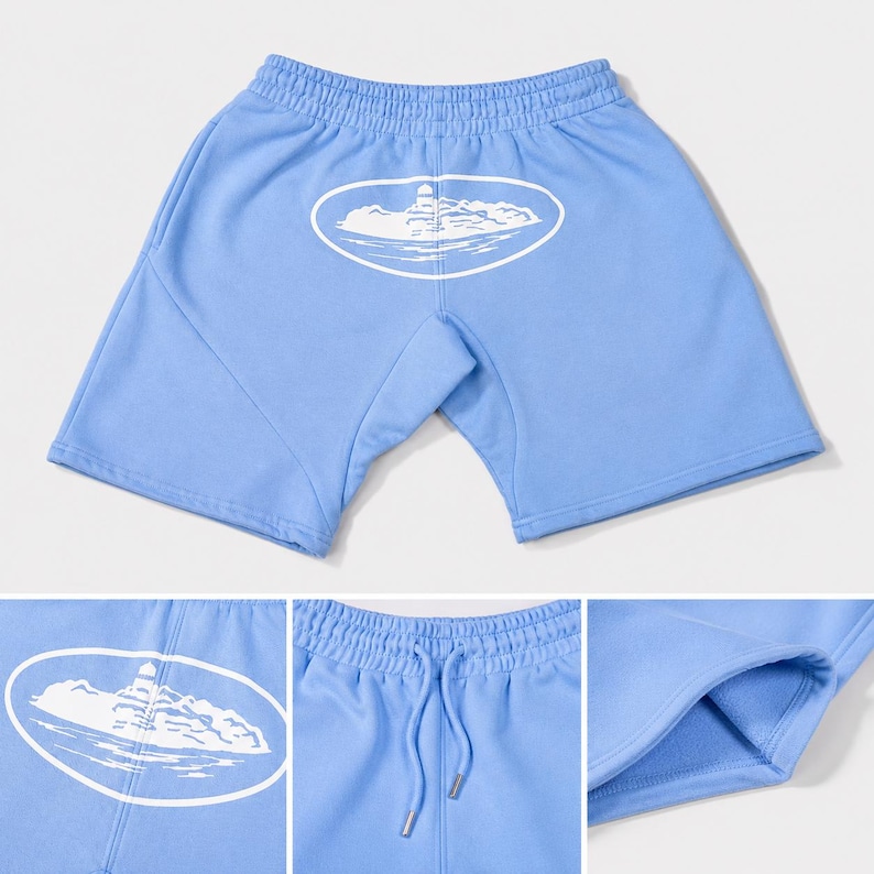 May include: Light blue athletic shorts featuring an elastic waistband with a drawstring and a white oval graphic on the front depicting a lighthouse on a rocky island. The shorts have a soft interior lining and visible side seam construction.