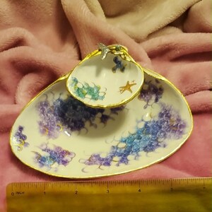 May include: A white ceramic two-tiered shell-shaped dish with gold-painted edges, featuring blue and purple scale patterns and a small starfish motif. A silver-toned mermaid tail ornament sits on the top tier. A ruler shows a width of 6 inches.