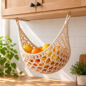 May include: A cream-colored macrame fruit hammock suspended from the underside of wooden kitchen cabinets by two metal hooks. The woven net holds a collection of bright orange citrus fruits, including oranges and lemons, within its mesh.