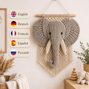May include: A macrame wall hanging featuring a three-dimensional grey elephant head with large, fringed ears and cream-coloured tusks. The elephant is centred on a cream-coloured woven macrame panel suspended from a horizontal wooden dowel.