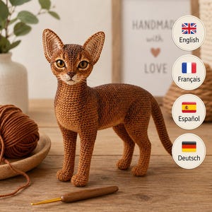 Peut inclure: Une figurine de chat abyssin crocheté de couleur brune avec de grandes oreilles pointues et des yeux ambrés. Le chat est représenté debout sur ses quatre pattes, avec une surface en fil texturé détaillée. Un crochet en bois est posé à côté.
