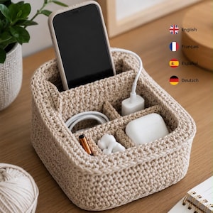 May include: A beige crocheted desk organizer with multiple compartments. It holds a smartphone, a white charging cable, a white power adapter, a white wireless earbud case, and a small white figurine. The organizer has a structured, woven texture.