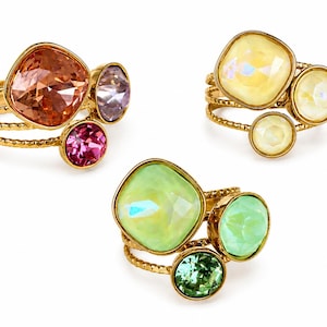 May include: Three gold-toned rings with textured bands, each featuring a cluster of three faceted crystals in varying sizes. The first ring has orange, purple, and pink stones; the second has yellow stones; the third has light and dark green stones.