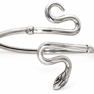 Peut inclure: Un bracelet en métal argenté poli en forme de serpent enroulé. Le design présente une bande lisse et continue qui s'enroule, se terminant par une tête de serpent détaillée à une extrémité et une queue incurvée et effilée à l'autre.