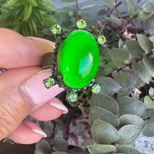 Peut inclure: Une bague ornée d'un grand cabochon ovale d'un vert citron éclatant, monté sur une structure en métal sombre. Le sertissage est agrémenté de six petits cristaux facettés vert clair disposés autour de la pierre centrale en rayons de soleil.