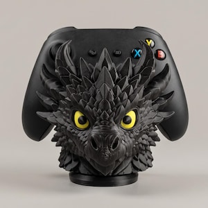 Könnte beinhalten: Ein schwarzer Gaming-Controller-Ständer mit einem detaillierten, skulptierten Drachenkopf mit markanten Schuppen und leuchtend gelben Augen. Der mattschwarze Drachenkopf ist mittig platziert, während der Controller auf einem runden Sockel dahinter ruht.